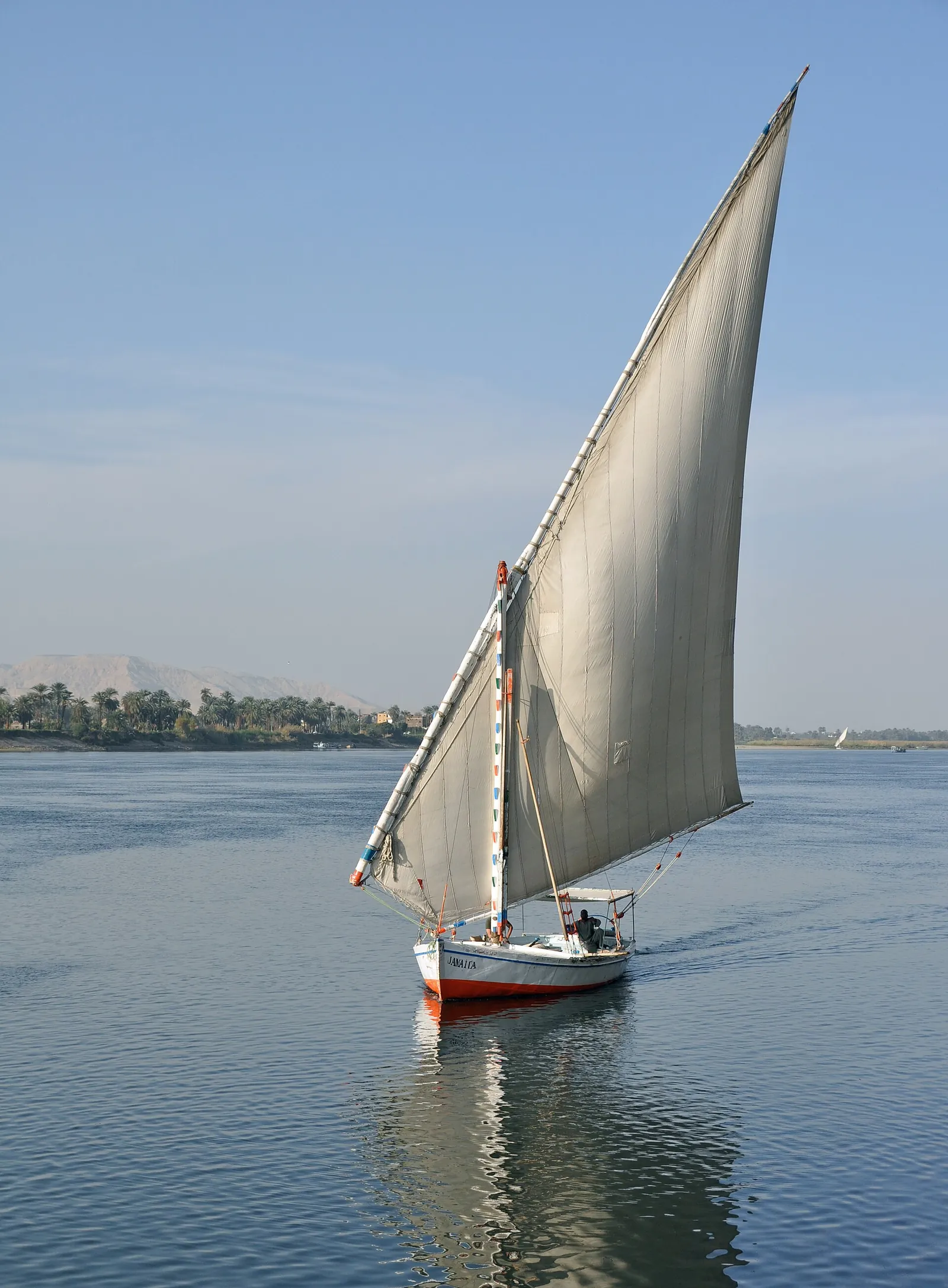 Felucca under full sail on the Nile with palm trees and desert hills on the far bank