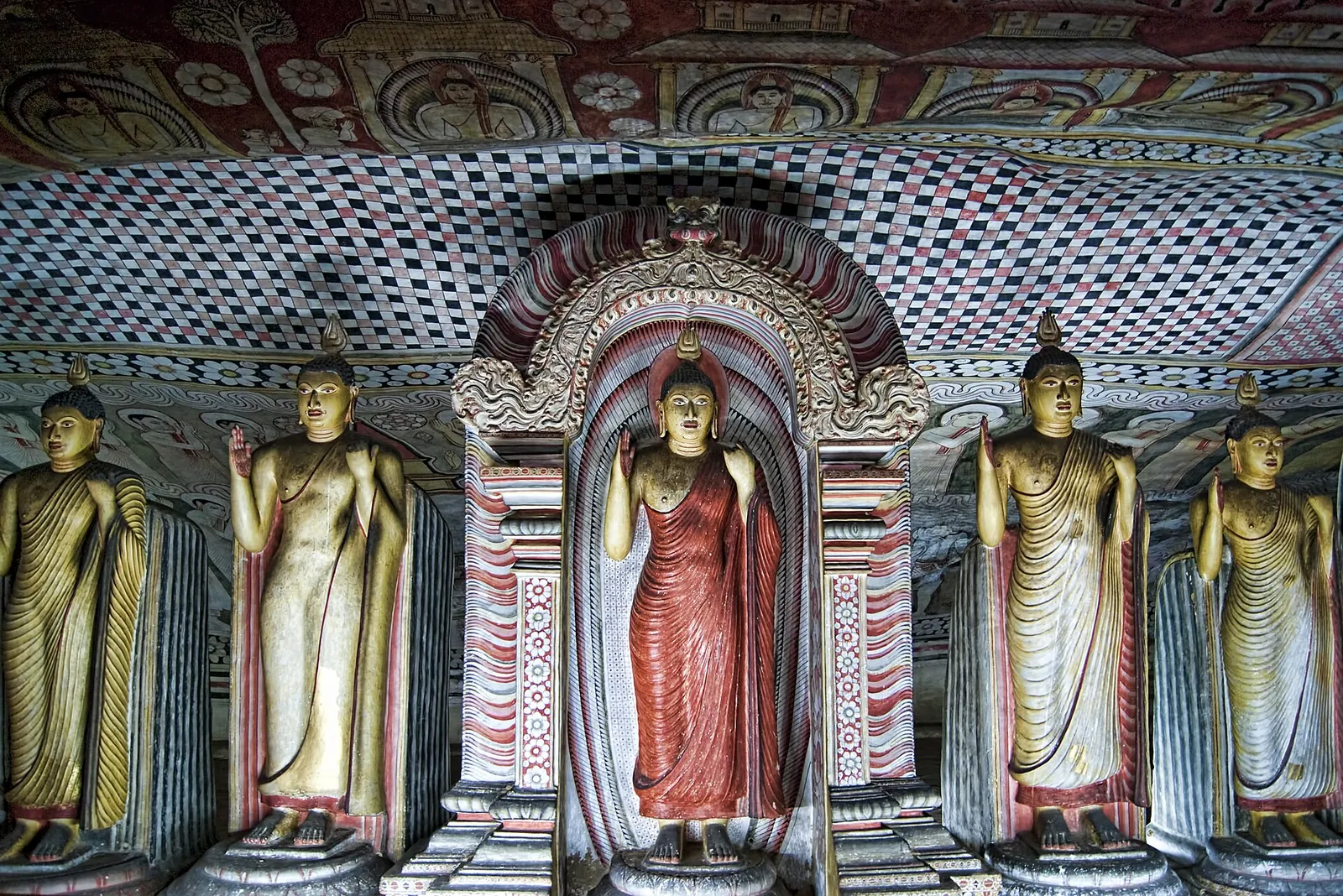 A row of painted Buddha statues inside the Dambulla cave temple under a decorated rock ceiling